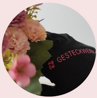 Das Bild zeigt einen schwarzen Arbeitshandschuh mit dem Firmenlogo von Gesteckwerk. Der Handschuh hält einen Trockenblumenstrauß in pink bis pfirsichfarben in der Hand und es wird im Hintergrund ein florales Design angefertigt.